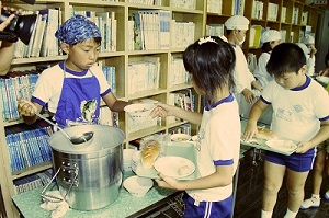 08強化磁器製の新食器を使用した配膳・白水小学校(平成11年8月、いわき市撮影)：少し重たい汁わんになり慎重に手渡しているように見えます