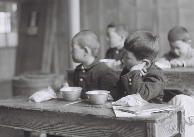 給食風景3・小玉小学校2昭和30年代、国府田英二氏提供)：脱脂粉乳といっしょに並んでいるのはクラッカーでしょうか？